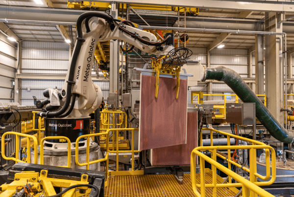 A robotic arm in a factory lifts large sheets with its gripper. Yellow railings and industrial equipment surround the workspace, and ducting is visible in the background.